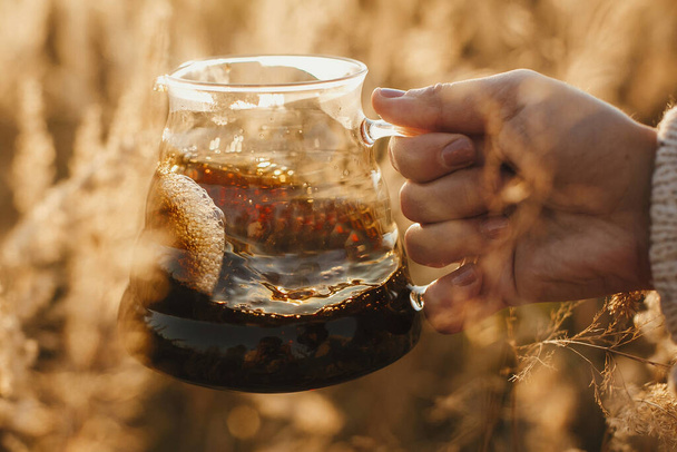 Podróżnik trzymający świeżą gorącą kawę w szklanej kolbie na tle słonecznego, ciepłego światła w wiejskich ziołach. Alternatywna kawa warząca się na świeżym powietrzu w podróży. Atmosferyczny, rustykalny moment spokoju. - Zdjęcie, obraz