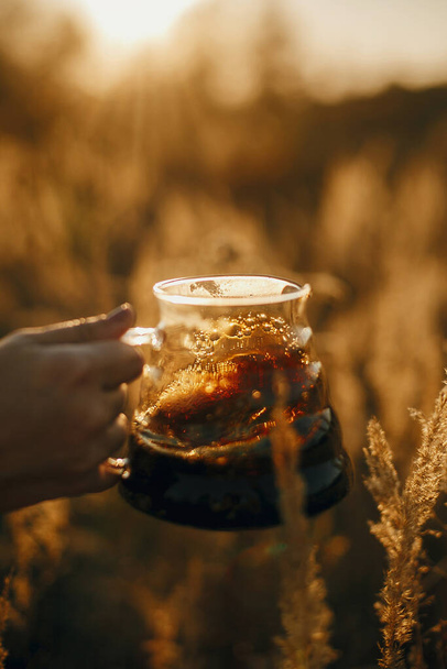 Alternative coffee brewing outdoors in travel. Hand holding fresh hot coffee in glass flask on background of sunny warm light in rural herbs. Atmospheric rustic tranquil moment. Vertical image - Foto, afbeelding