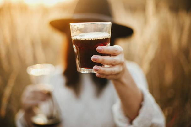 Hipster woman holding hot coffee in glass cup on background of rural herbs in sunset. Alternative coffee brewing outdoors in sunny light. Atmospheric rustic moment. Travel and Wanderlust - Foto, imagen