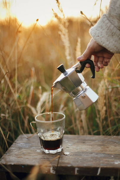 Hipster pouring fresh hot coffee from geyser coffee maker into glass cup in sunny warm light in rural countryside herbs. Atmospheric tranquil moment. Alternative coffee brewing in travel - Valokuva, kuva