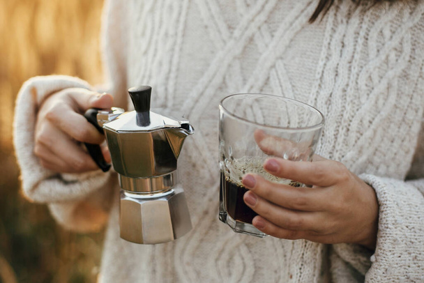 Hipster γυναίκα κρατώντας geyser καφετιέρα και ποτήρι με καφέ σε ηλιόλουστο ζεστό φως στην ύπαιθρο βότανα. Ατμοσφαιρική ρουστίκ στιγμή. Εναλλακτική παρασκευή καφέ σε ταξίδια. - Φωτογραφία, εικόνα