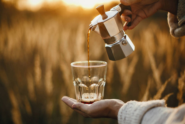 Альтернативный кофе для путешествий. Залить свежий горячий кофе из гейзер кофеварка в стеклянную чашку в солнечном теплом свете в сельской местности травы. Атмосферный момент. Вертикальное изображение
 - Фото, изображение