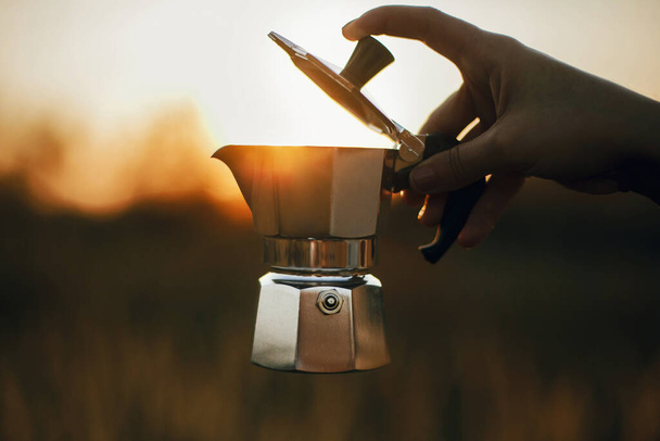 Мандрівник тримає кавоварку Гейзера зі свіжою кавою в сонячному теплому світлі в сільській місцевості. Атмосферний сільський момент. Альтернативне приготування кави у подорожах
. - Фото, зображення