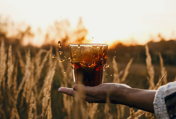 Coffee splash and drops in sun. Spilling hot coffee from glass cup on hand in sunny warm light in countryside rural herbs. Alternative coffee brewing in travel. Atmospheric tranquil moment. - Photo, Image