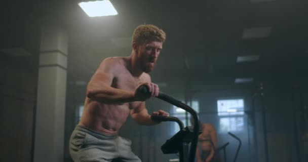 a hardy man overcoming fatigue trains on a stationary bike in a fitness room. atmospheric gym. relief athlete - Imágenes, Vídeo