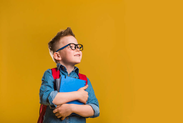 cuki kisfiú szemüvegben, könyvvel a kezében. gyerek piros táska elszigetelt sárga. - Fotó, kép