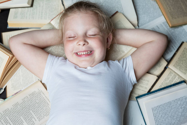 A smiling little girl lies with closed eyes on a pile of open books. Top view. Education and training. Close-up. - Fotoğraf, Görsel