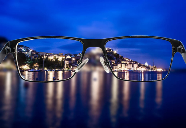 Fokussierter nächtlicher Stadtblick beim Blick durch den Brillenrahmen. Schwarze Metallbrillen blicken durch das bunte Stadtbild. Brille für Menschen mit Sehbehinderung. Gesundheitszustand - Foto, Bild