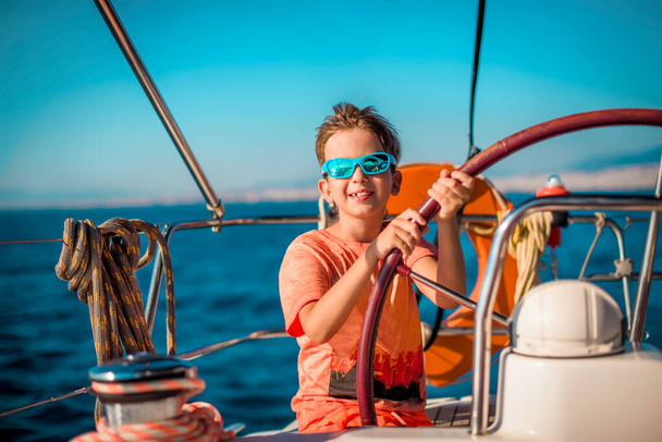 Pohledný chlapec ve věku 9-10 let u kormidla jachty v jasně oranžových šatech, s modrými brýlemi proti modré obloze. Námořní dovolená koncept, plachtění regata. Cestování s dětmi. - Fotografie, Obrázek