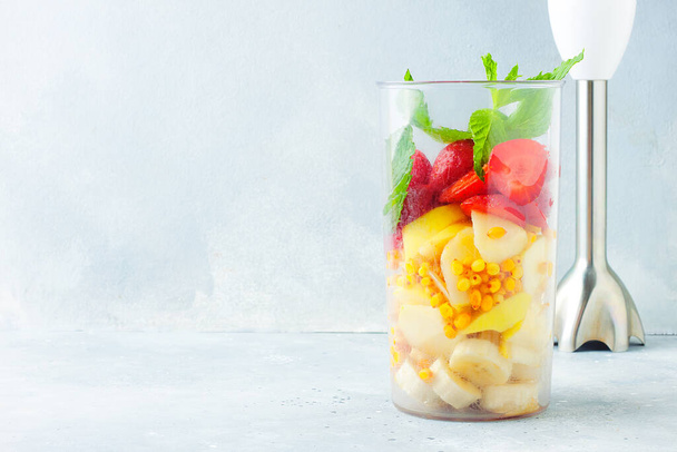 Blender filled with different fruits for vitamin smoothie - banana, strawberry, apple, sea-buckthorn and mint. Grey stone background, copy space. Healthy eating concept - Photo, Image
