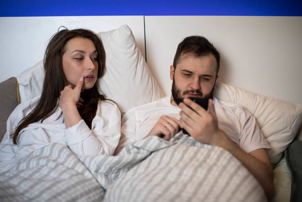 Молодая супружеская пара лежит в постели, мужчина использует мобильный телефон, игнорируя свою жену
 - Фото, изображение