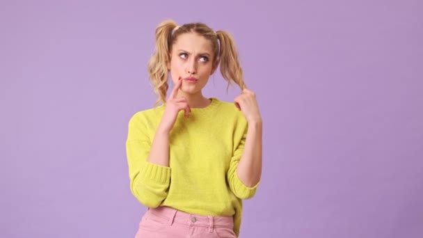 An unrivaled blonde girl thoughtfully puts hair on her finger, shows the index finger of her hand near her cheek in an isolated studio on a purple background - 映像、動画