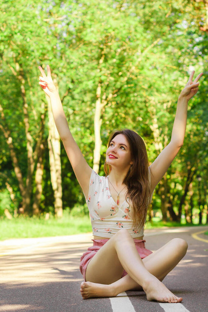 Girl in pink shorts and white top smiling and sitting in the middle of the road in the park with legs crossed and hands up in victory sign. Green grass and trees around. End of pandemia and quarantine - Foto, afbeelding