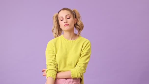 A great blonde girl in a yellow sweater is bored, standing down with her hands down in an isolated studio on a purple background - Filmagem, Vídeo