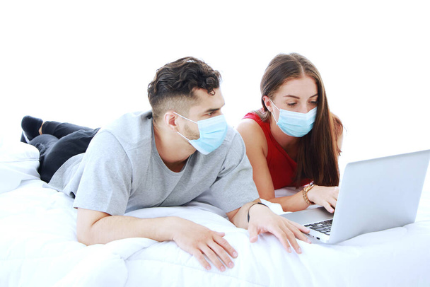 Maske takan beyaz bir çift yatak odasındaki sosyal medya teknolojisinde covid-19 virüsünün dizüstü bilgisayarla rahatlamasını engelliyor. Tatilde evli aile hayatı için sosyal mesafeler ve sağlık hizmetleri. - Fotoğraf, Görsel