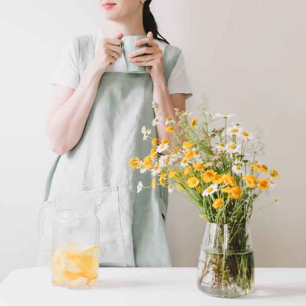 Woman in linen apron drinking lemonade with cozy home background. Clean natural fabric apron. Food blogger, homemade cooking - Photo, Image