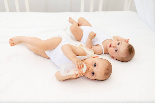 dos niños un niño y una niña gemelas de 8 meses beben leche de un biberón en la cama en el vivero, alimentando al bebé, concepto de comida para bebés, vista superior, lugar para el texto
 - Foto, imagen