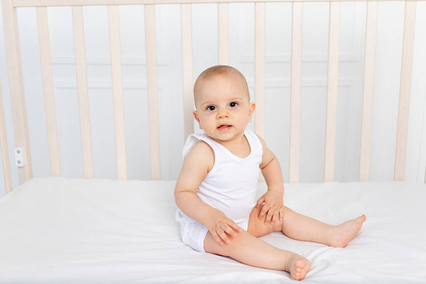 Baby Junge 8 Monate alt sitzt in einem Kinderbett in einem Kinderzimmer in weißen Kleidern und blickt in die Kamera, der Morgen des Babys - Foto, Bild