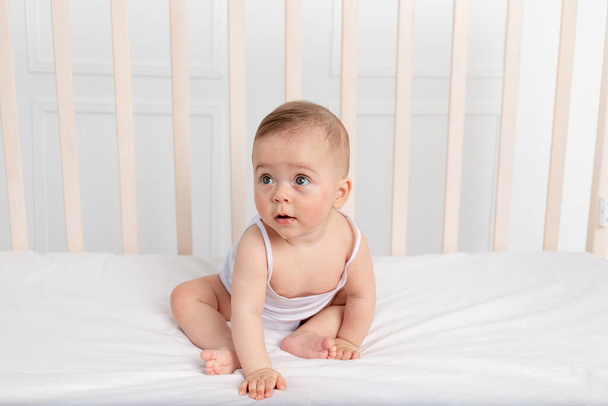 χαμογελαστό κοριτσάκι 8 μηνών που κάθεται σε μια κούνια σε παιδικό δωμάτιο με λευκά ρούχα και φιόγκο και κοιτάζει μακριά, το μωρό πρωί, το μωρό έννοια προϊόντα μωρό - Φωτογραφία, εικόνα