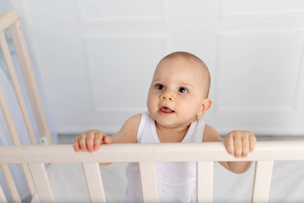 muotokuva hymyilevä vauva poika 8 kuukautta vanha seisoo pinnasänky lastenhuoneessa valkoisissa vaatteissa ja katselee pois, aamu vauva, vauvan tuotteet käsite - Valokuva, kuva