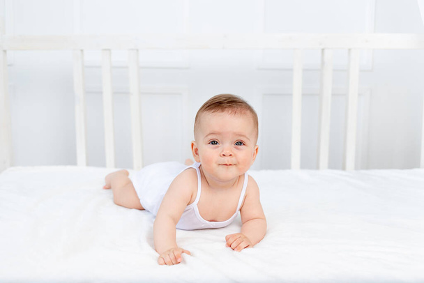 niña sonriente de 6 meses acostada en la cama en el vivero sobre su estómago y mirando a la cámara, bebé de la mañana, concepto de productos para bebés
 - Foto, imagen