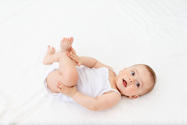 kislány 6 hónapos feküdt egy bölcsőben a gyerekszobában a hátán, és fogta a lábát, a kamerába nézett, baba reggel, baba termékek koncepció - Fotó, kép