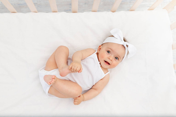 6 Monate altes Mädchen liegt mit weißer Kleidung auf dem Rücken in einem Kinderbett im Kinderzimmer und lacht, blickt in die Kamera, Babymorgen, Babyprodukte-Konzept - Foto, Bild