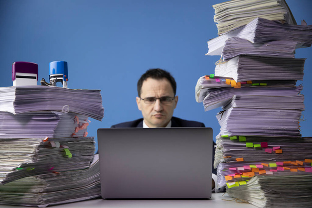 hombre de negocios trabaja en el ordenador portátil en el escritorio de la oficina con una gran pila de documentos
 - Foto, Imagen