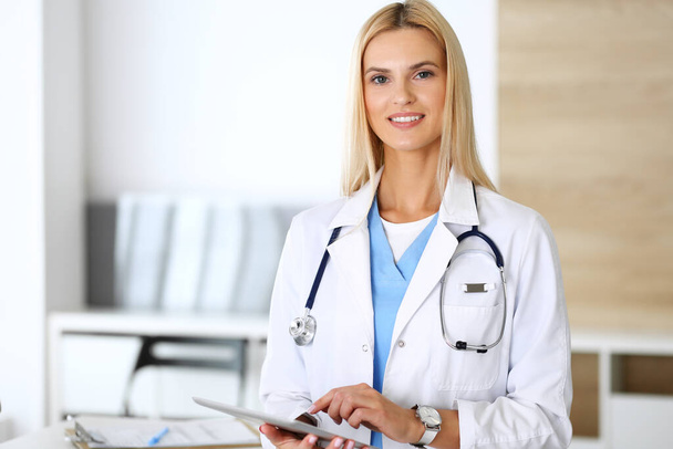 Doctor woman at work in hospital excited and happy of her profession. Blonde physician controls medication history records and exam results while using tablet computer. Medicine and healthcare concept - Photo, image