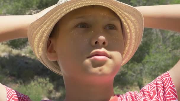 Κοντινό πορτραίτο αγοριού με καπέλο την ηλιόλουστη καλοκαιρινή μέρα. Παιδί σηκώνοντας τα χέρια και κοιτάζοντας γύρω από την εξωτερική - Πλάνα, βίντεο