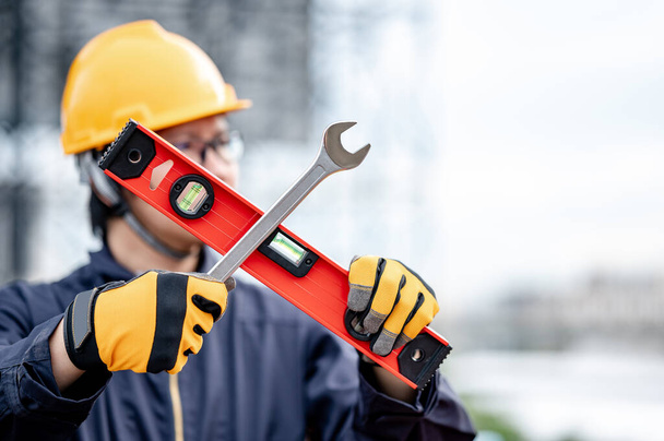 Мужчина азиатский механик или обслуживающий работник в защитном костюме и шлеме, держащий гаечный ключ и инструмент в крестообразной форме на строительной площадке. Оборудование для машиностроения - Фото, изображение