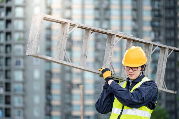 Ein asiatischer Bauarbeiter mit Schutzhelm und grüner Weste trägt eine Alu-Leiter auf der Baustelle. Bauingenieurwesen, Architekturbau- und Haustechnikkonzepte - Foto, Bild