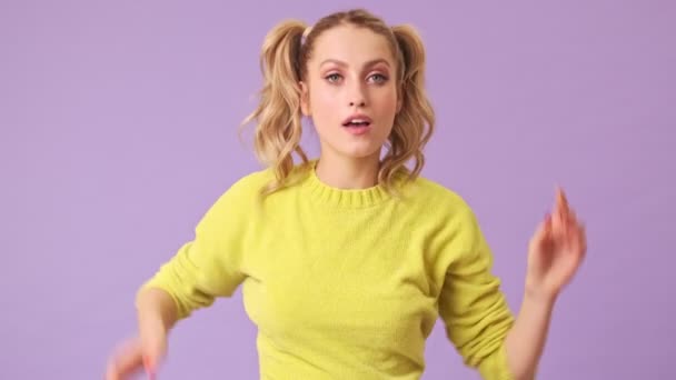A charming blonde in a yellow sweater prepares for a date, straightens her ponytails on her head, takes out a makeup brush and begins to paint her face in an isolated studio on a purple background - Metraje, vídeo