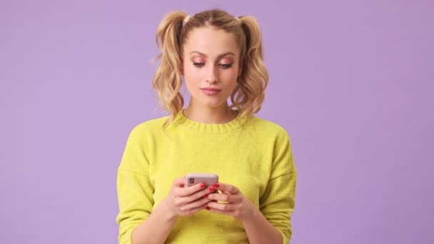 A magical blonde girl writes a message on her phone in an isolated studio on a purple background - Séquence, vidéo