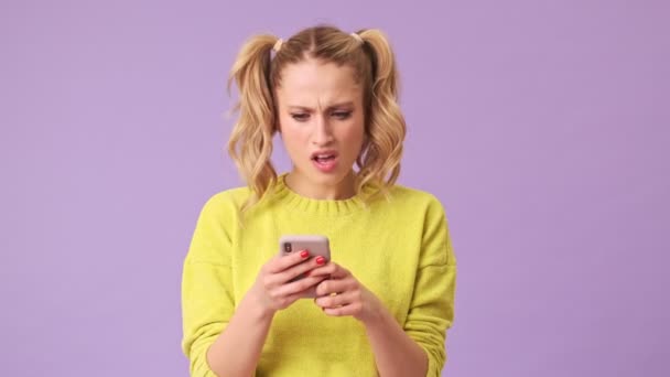 A cute blonde writes on the phone an answer to the interlocutor to whom she is unpleasantly surprised in an isolated studio on a purple background - Séquence, vidéo