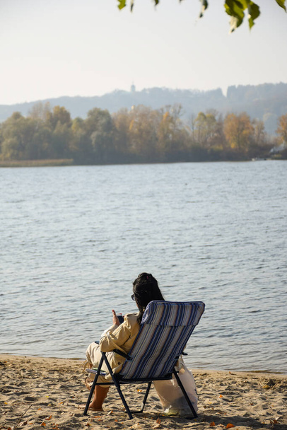 La chica con el pelo negro se sienta en la orilla del río en la arena en una tumbona y mira el teléfono móvil
 - Foto, Imagen