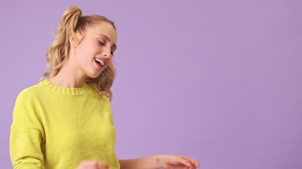 Charming blonde dancing with a smile, twisting hair on her fingers in an isolated studio on a purple background - Materiaali, video