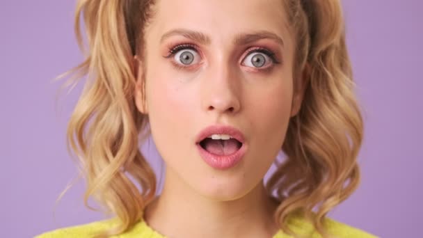 Portrait of a beautiful blonde with a surprised face and open mouth in isolated studio on a purple background - Imágenes, Vídeo