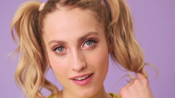 Portrait of a beautiful young smiling blonde flirting at the camera, biting her lip and twisting hair over her finger in an isolated studio on a purple background - Filmati, video