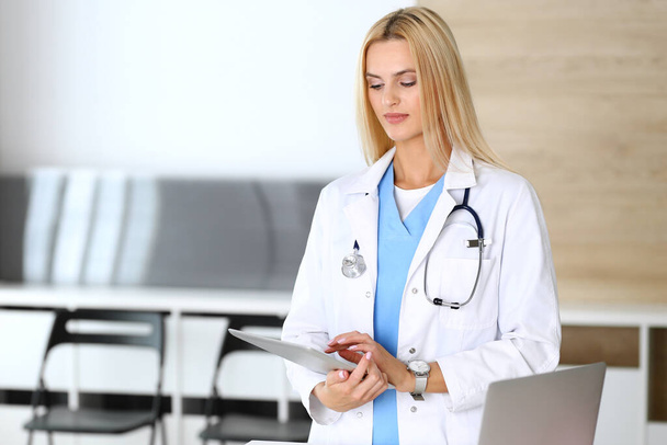 Doctor woman at work in hospital excited and happy of her profession. Blonde physician controls medication history records and exam results while using tablet computer. Medicine and healthcare concept - Photo, image