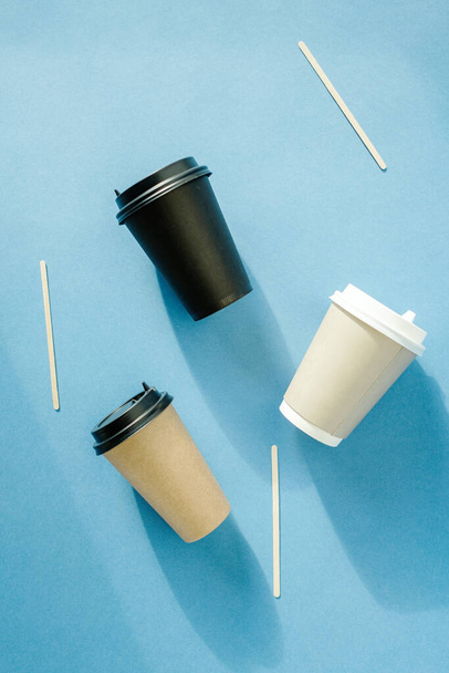 Three multi-colored paper cups for coffee and wooden sticks for mixing sugar lie on a blue background. Coffee cups with hard shadows. Takeaway drinks concept.Template for design. - Photo, Image