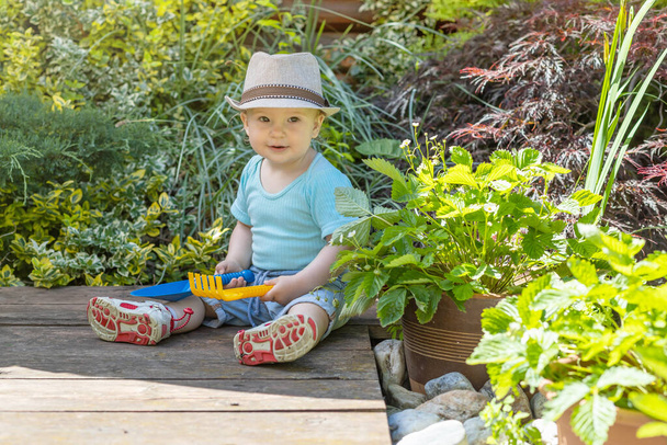 Χαριτωμένο αγοράκι παίζει με το μωρό τσουγκράνα και φτυάρι κάθεται στον κήπο - Φωτογραφία, εικόνα