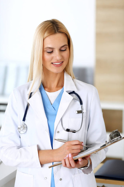 Doctor woman at work in hospital excited and happy of her profession. Blonde physician controls medication history records and exam results while using clipboard. Medicine and healthcare concept - 写真・画像