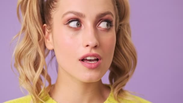 A cute blonde looks around in order not to divulge a secret, and shows silence gesture in an isolated studio on a purple background - Imágenes, Vídeo