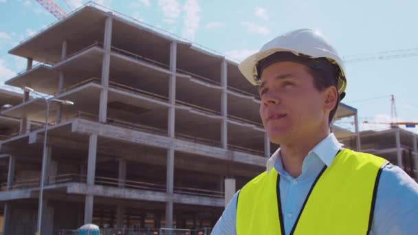 Professional builder standing in front of construction site. Foreman in hardhat helmet and vest. Office building and crane background. Business, real estate and investment concepts. - Séquence, vidéo