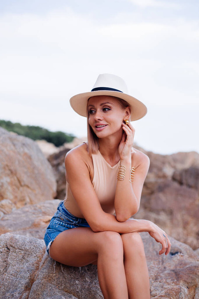 Зовнішній модний портрет молодої жінки євро в бежевому костюмі, сині джинсові шорти і класичний стильний капелюх на тропічному скелястому пляжі, теплий захід сонця.. - Фото, зображення