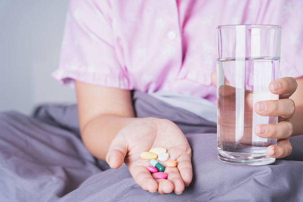 Κοντινό πλάνο γυναίκα ασθενής κρατώντας ένα ποτήρι νερό και χάπια φάρμακα στην κρεβατοκάμαρα. Έννοια υγειονομικής και ιατρικής φαρμακευτικής - Φωτογραφία, εικόνα