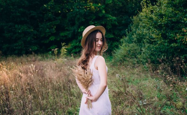 Kaunis tyttö, jolla on kiharat hiukset hatussa. Brunette tyttö kävelee alalla kukkia. Nuori nainen kesällä kukkakimpun kanssa - Valokuva, kuva