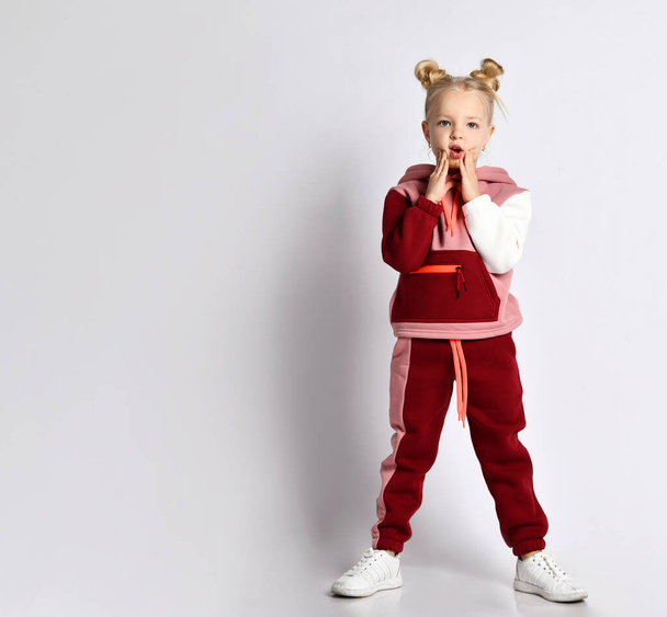 Little blonde child with buns hairstyle, in colorful sport suit, sneakers. She touching her cheeks, posing isolated on white - Foto, Imagem