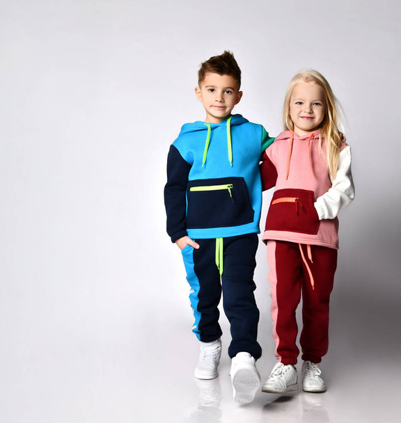 Little boy and girl in bright sportsuits walking forward hand in hand. Full length portrait isolated on light grey, copy space - Fotografie, Obrázek